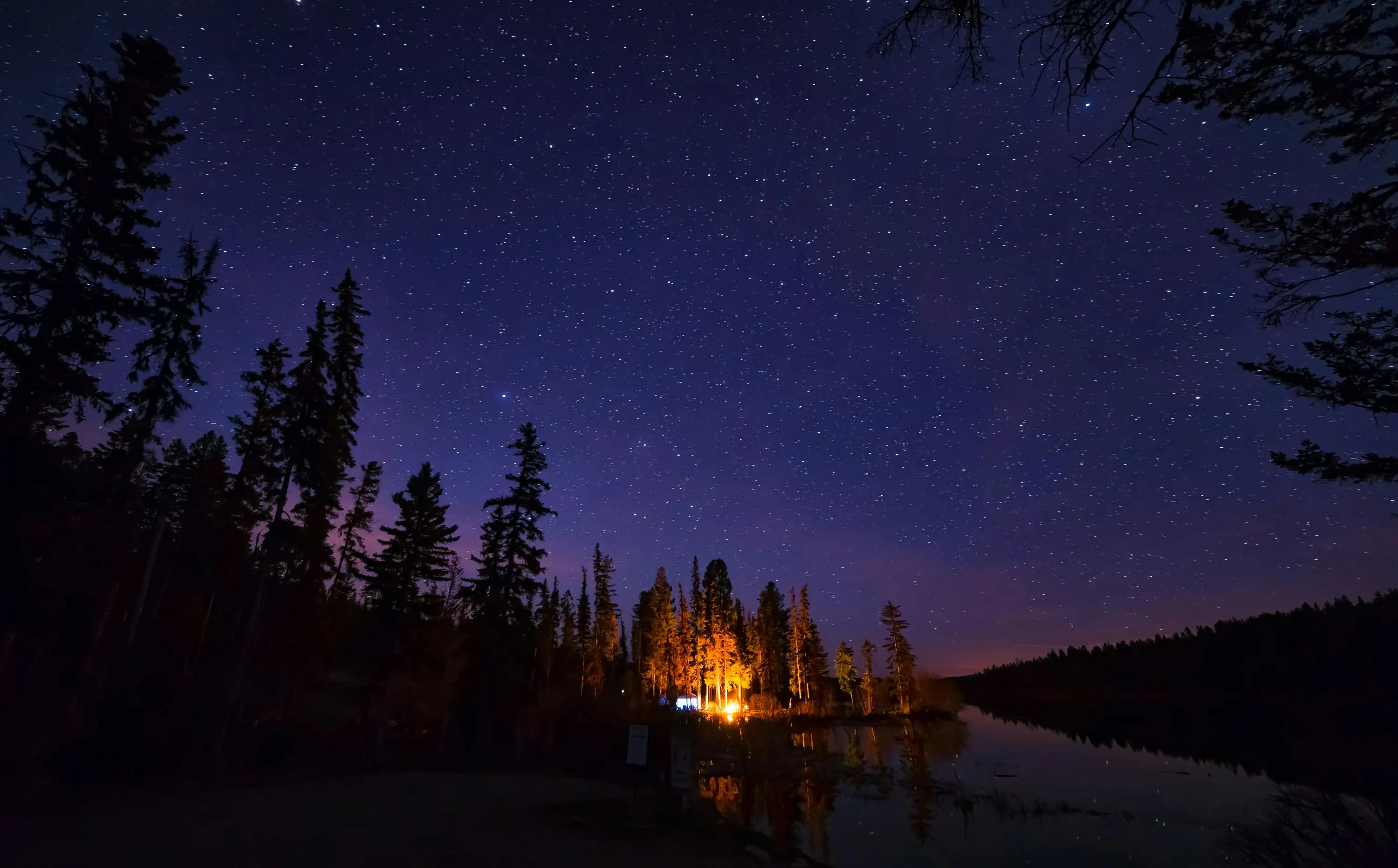 A starry view of a forest and lake at night. There is a campsite and campfire visible on the other side of the lake.